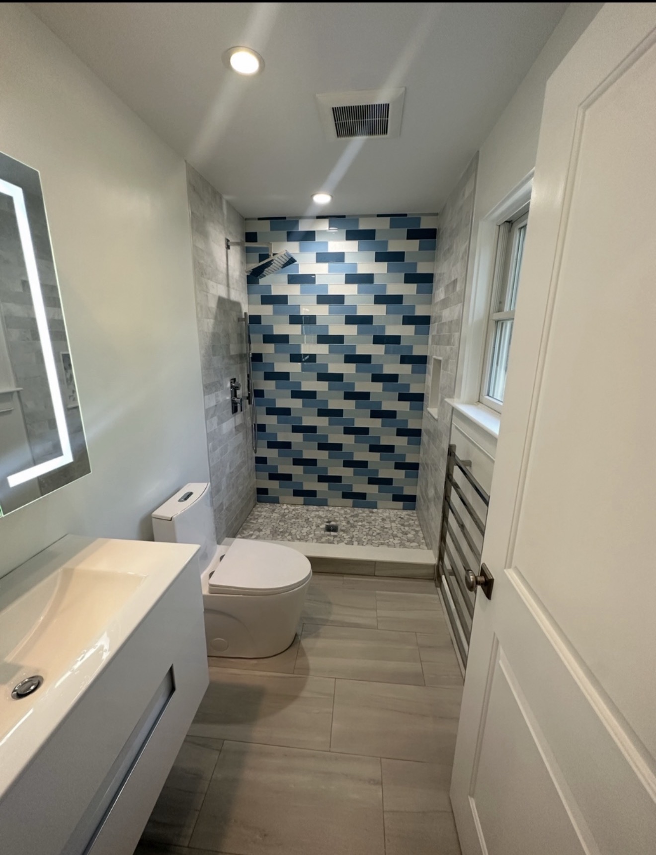 Modern walk-in shower bathroom remodel with stacked blue and white glass mosaic accent wall, gray porcelain tile, pebble shower floor, floating vanity, and LED mirror
