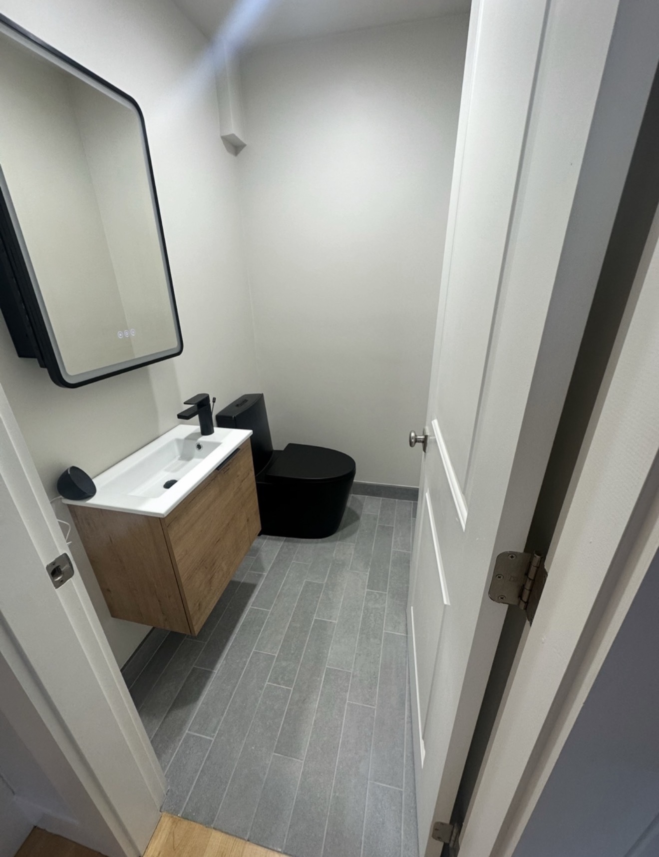 Modern powder room remodel with floating wood-grain vanity, matte black faucet, wall-hung black toilet, LED mirror, and gray plank tile floor